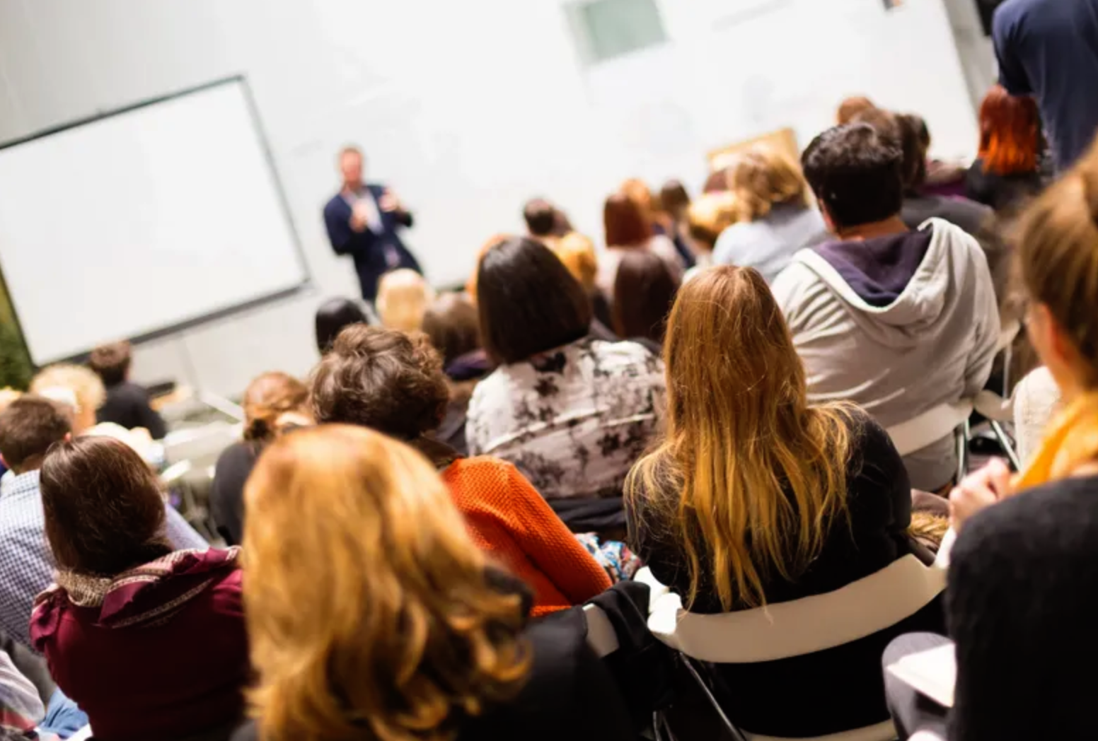 An audience seated and listening to a speaker presenting at the front of a conference room.