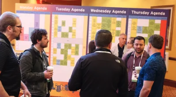 A group of people chat with coffee in front of color-coded conference agenda boards for the week.