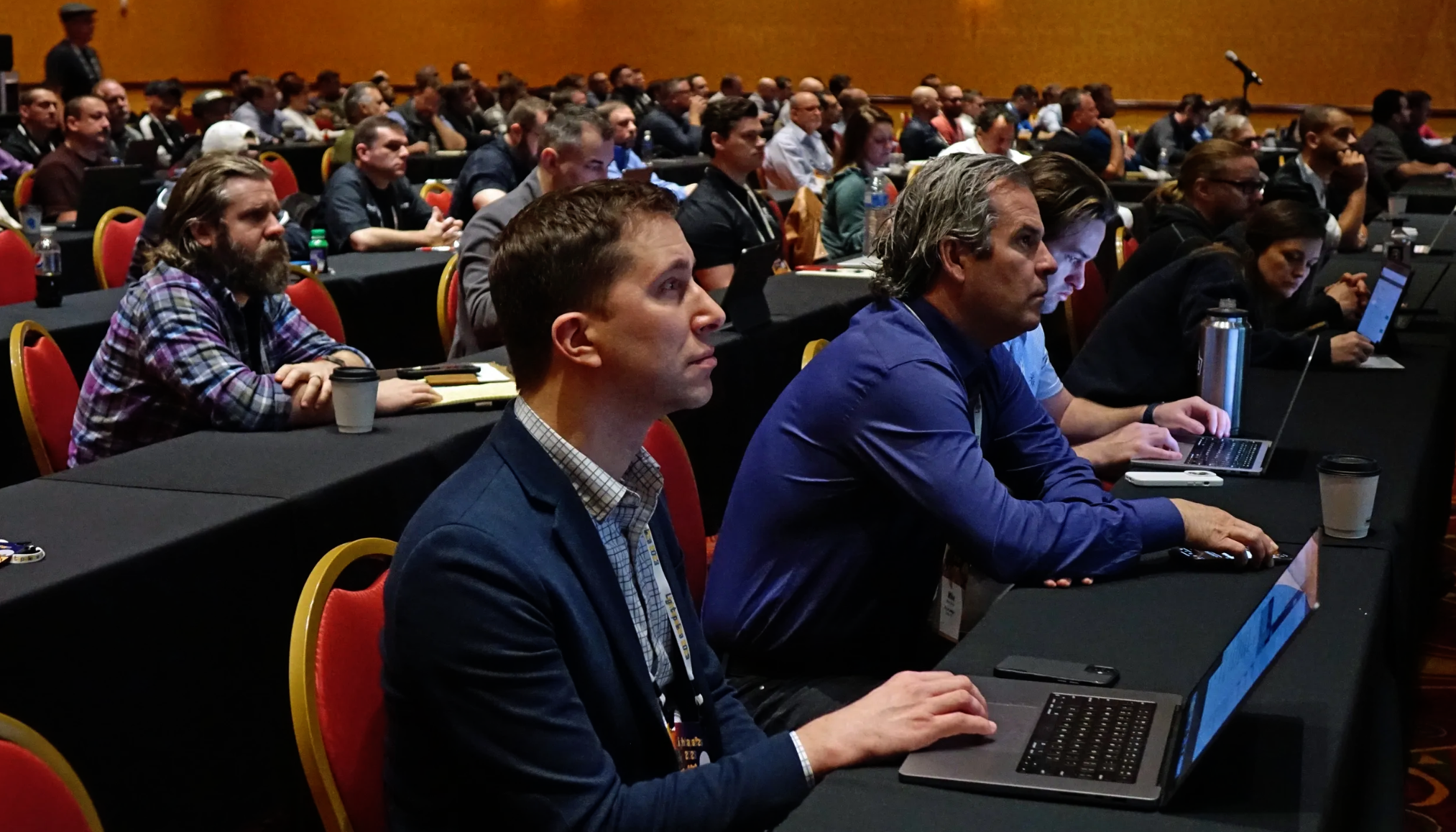 Conference attendees seated at long tables, listening and taking notes on laptops.